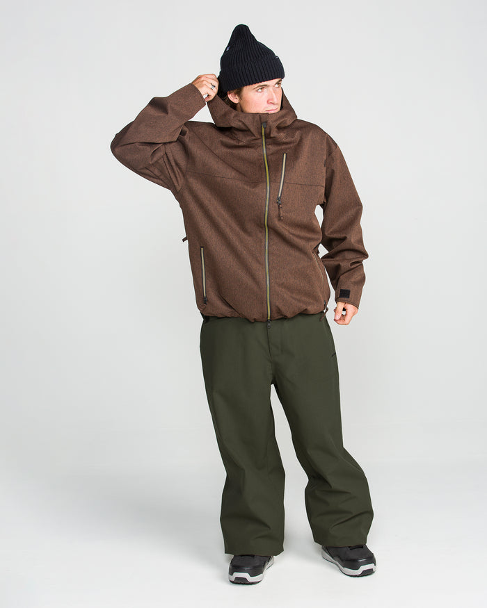 A person wearing a brown jacket, Volcom Men’s Lapper Pants in olive green, a black knit hat, and black shoes stands against a plain white background, adjusting their collar with one hand.