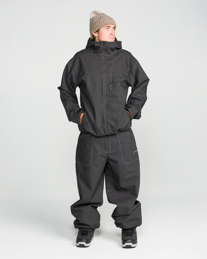 A person wearing Volcom Men’s Lapper Pants—baggy dark gray 10K waterproof pants—paired with a matching hooded jacket, hands in pockets, a beige knitted hat, and black sneakers, standing against a plain white background.