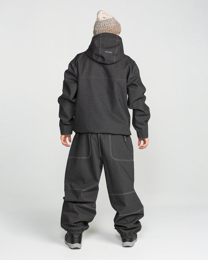 A person stands facing away, wearing a black hooded jacket, Volcom Men’s Lapper Pants in black, black shoes, and a beige knit beanie against a plain white background.
