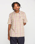 A man with short dreadlocks models the Volcom Men's Ledges Short Sleeve Shirt, featuring a classic fit and beige, pink, and white yarn-dyed plaid pattern, paired with dark jeans and set against a plain white background.