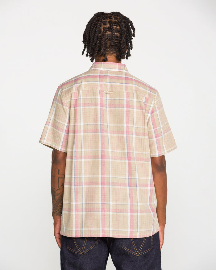 A person with short, twisted hair and visible tattoos on their left arm stands with their back to the camera, wearing the Volcom Men's Ledges Short Sleeve Shirt—a classic-fit, beige yarn-dyed plaid shirt with pink and white stripes—paired with dark jeans.