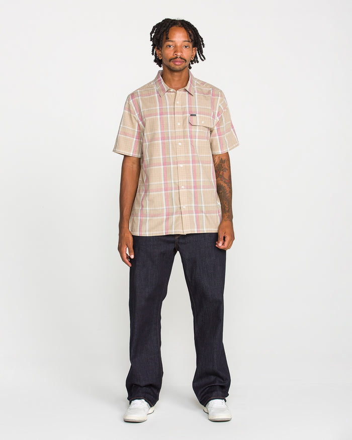 A man stands facing forward, wearing the Volcom Men's Ledges Short Sleeve Shirt in a classic-fit yarn-dyed plaid, paired with dark jeans and white sneakers against a plain white background.