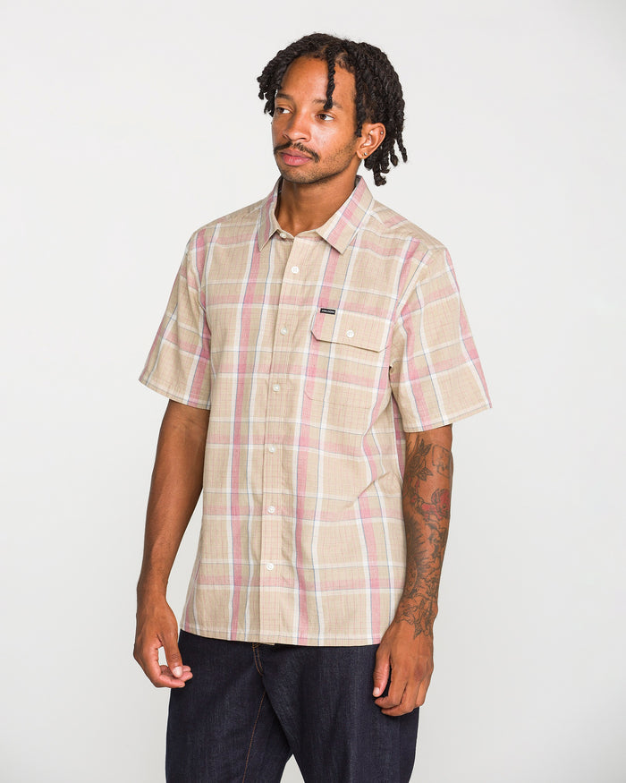 A man with short dreadlocks wears the Volcom Men's Ledges Short Sleeve Shirt, a yarn-dyed plaid classic-fit button-up featuring a front pocket, while standing against a plain white background.