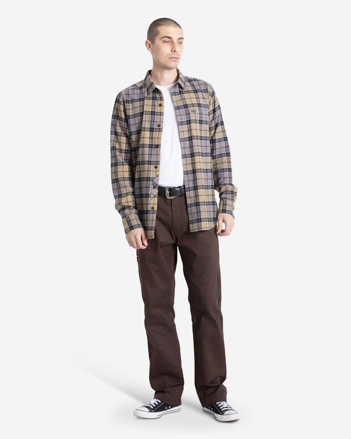 A man with short hair stands facing slightly right in a Volcom Men's Caden Plaid Long Sleeve shirt over a white tee, brown pants, black belt, and black-and-white sneakers. He has a neutral expression against a plain white background.