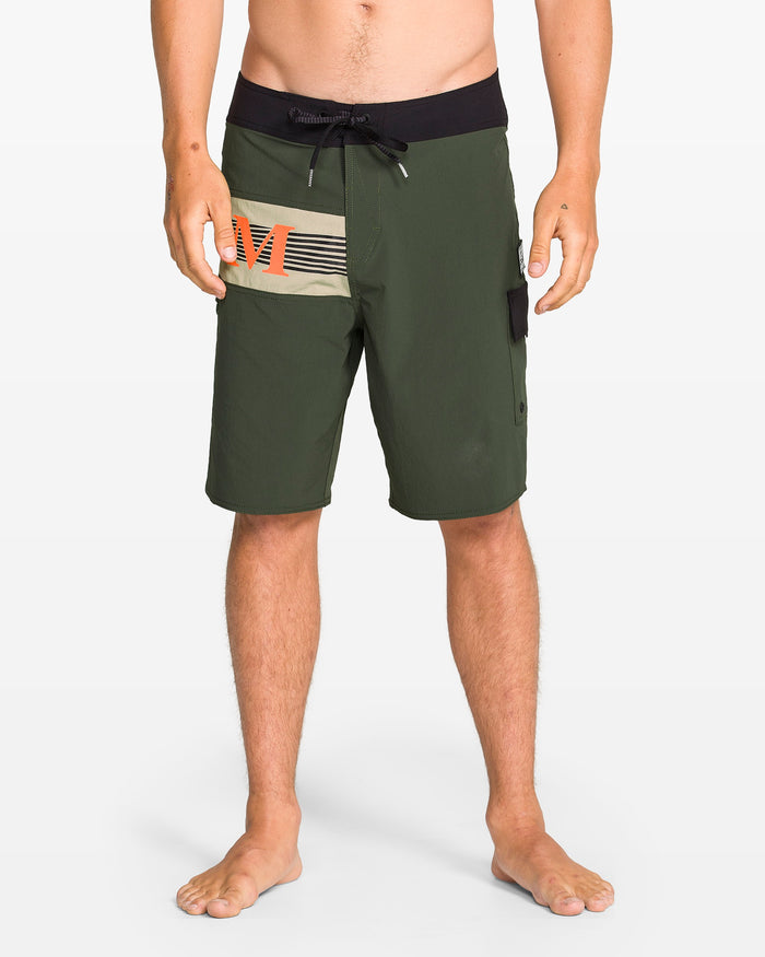 A person stands barefoot in Volcom Men's Liberator Mod olive green 20" board shorts with 4-way stretch nylon, black waistband with Cinch Fly Technology, drawstring, side pocket, and beige/orange stripes on the left thigh.