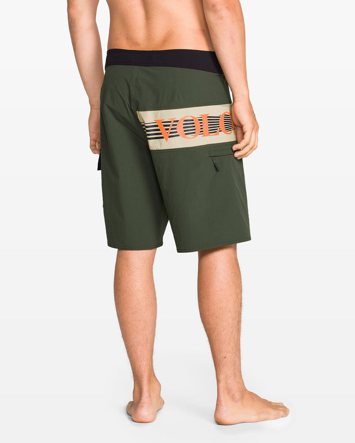 A man stands barefoot, wearing Volcom Men's Liberator Mod olive green 20" board shorts with Cinch Fly Technology, a black waistband, and a large side pocket featuring bold orange and black "VOLCOM" lettering on a white background.