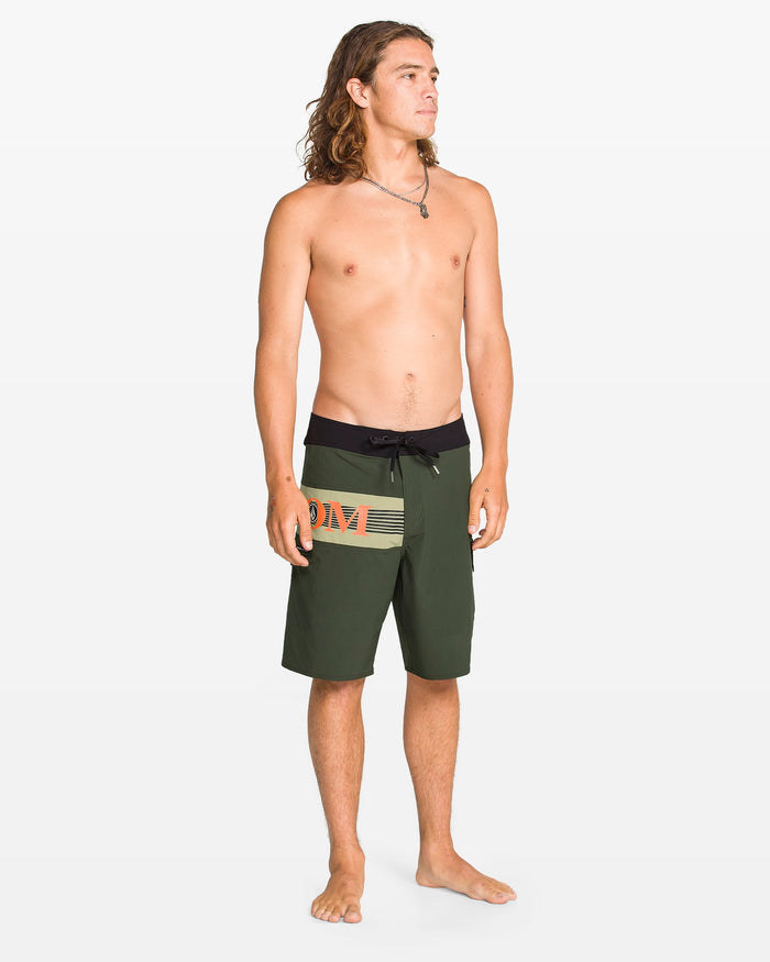 A young man with long hair stands barefoot against a white background, wearing Volcom Men's Liberator Mod board shorts in dark green and black with orange and beige graphics. He is shirtless, looking slightly left, and features Cinch Fly Technology.