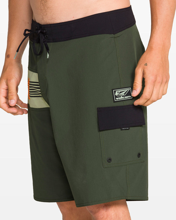 A person wears Volcom Men's Liberator Mod board shorts in dark green with a black waistband and pocket, logo patch, drawstring waist, and 4-way stretch nylon. Only the torso and upper legs are shown.