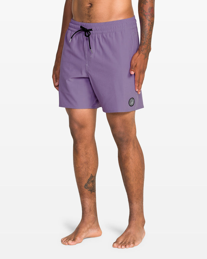 A barefoot person wears Volcom Men's Lido Solid 16" Trunk swimwear in light purple, featuring a black drawstring and circular patch on the left leg. Made from recycled polyester. The background is plain white.