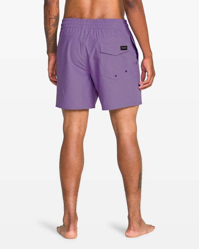 A person stands barefoot, facing away, in Volcom Men's Lido Solid 16" Trunk—purple recycled-polyester swimwear with a back pocket and black logo patch. A tattoo appears on their right arm. The background is plain white.
