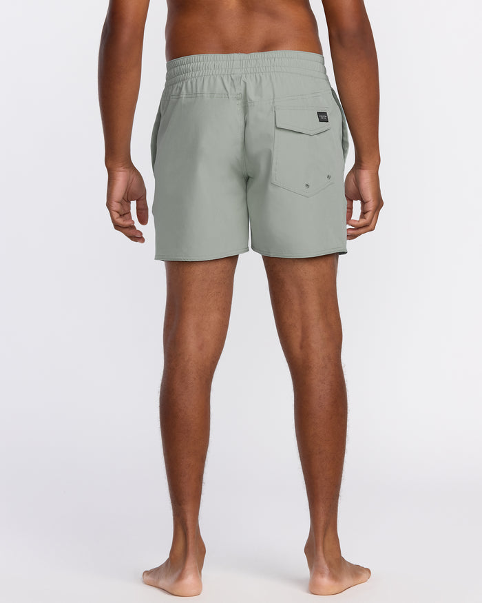 A barefoot person faces away, wearing Volcom Men's Lido Solid 16" Trunks in light gray with an elastic waistband and a right-side flap pocket, made from recycled polyester. The background is plain white.
