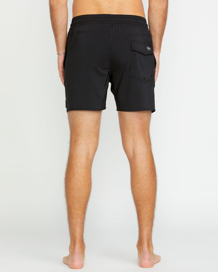 A barefoot person stands with their back to the camera, wearing Volcom Men's Lido 2 Solid Trunks—short black swim trunks made from recycled polyester, featuring an elastic waistband and a single flap pocket on the right side.