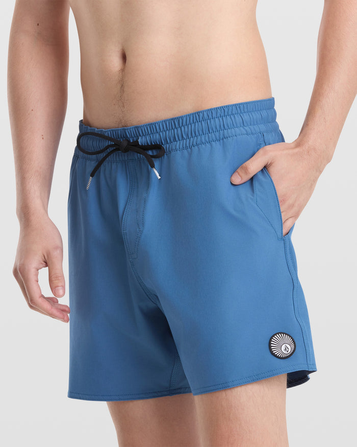 A person wears Volcom Men's Lido Solid 16" Trunks—blue with a black drawstring, made from recycled polyester, and featuring a circular white-and-black logo on the left leg—standing against a plain light background.