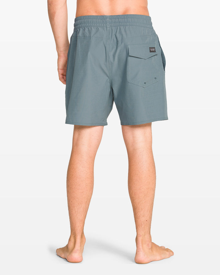 A person stands barefoot facing away, wearing Volcom Men's Lido Solid 16" Trunk boardshorts in teal with a 5-inch inseam, elastic waist, right flap pocket, and a small black label above the pocket. Background is plain white.