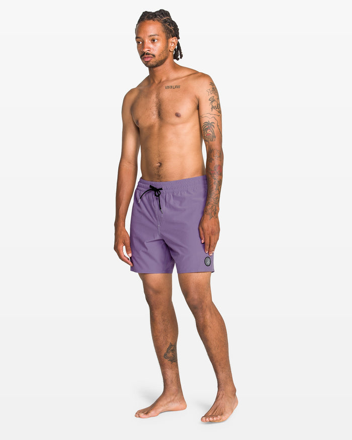 A tattooed man stands barefoot against a plain background, wearing Volcom Men's Lido Solid 16" Trunk in purple, made from recycled polyester with a black drawstring and a small round patch on the left leg.