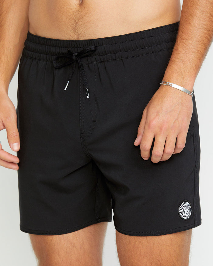 A person wears Volcom Men's Lido 2 Solid Trunks—black swim shorts made from recycled polyester, featuring a small circular white logo near the left hem, an elastic drawstring waistband, and side pockets. One hand rests relaxed by their side.