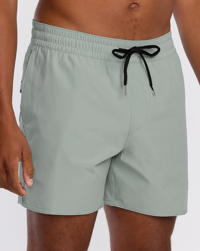 A person wears Volcom Men's Lido Solid 16" Trunks in light green, made from recycled polyester with 2-way stretch. The image shows only the lower torso and upper legs, set against a plain white background.