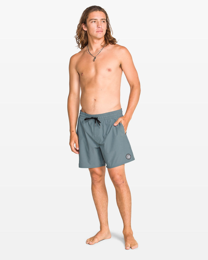 A young man with long brown hair, barefoot and wearing Volcom Men's Lido Solid 16" Trunk made from recycled polyester, stands against a plain white background, looking slightly to the side with one hand in his pocket.
