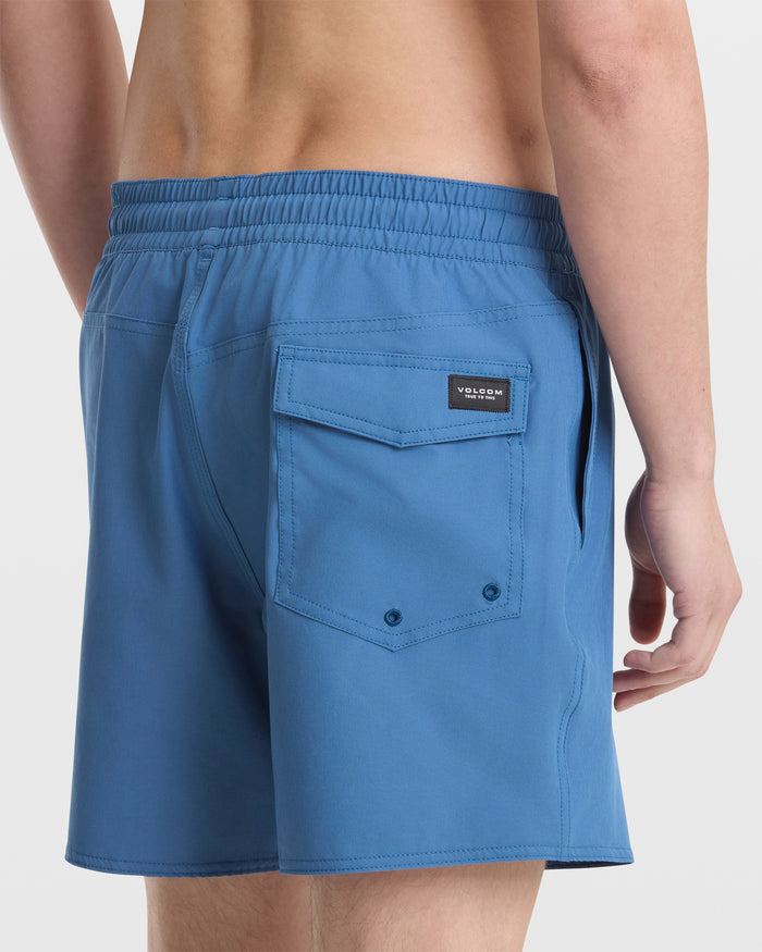 A person is shown from the waist down and behind, wearing Volcom Men's Lido Solid 16" Trunks in light blue, featuring a 16-inch outseam, elastic waistband, back flap pocket, and a small black logo tag.