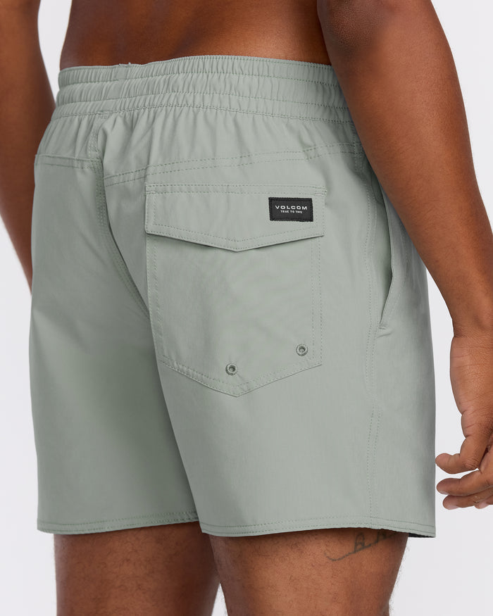 A person wears light gray Volcom Men's Lido Solid 16" Trunks with an elastic waistband, back pocket with flap, and black brand label. Made from recycled polyester 2-way stretch, these trunks are shown from the back and side.