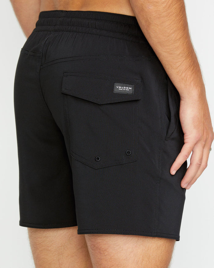 A person is shown from the back wearing black Volcom Men's Lido 2 Solid Trunks made from recycled polyester, highlighting a right-side flap pocket with a Volcom label. Their right hand rests by their side.