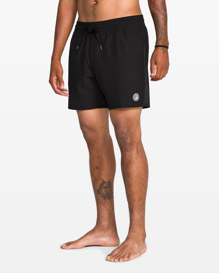 A man with tattoos on his forearm and lower leg stands barefoot against a plain white background, wearing Volcom Men's Lido 2 Solid Trunks—black swim trunks made from recycled polyester with mechanical stretch and a small circular logo.