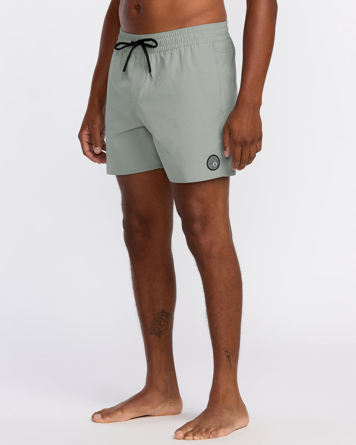 A person stands barefoot in Volcom Men's Lido Solid 16" Trunks, light gray with a black drawstring and small circular logo on the left leg. Made from recycled polyester with 2-way stretch. Torso and head not visible. Plain white background.