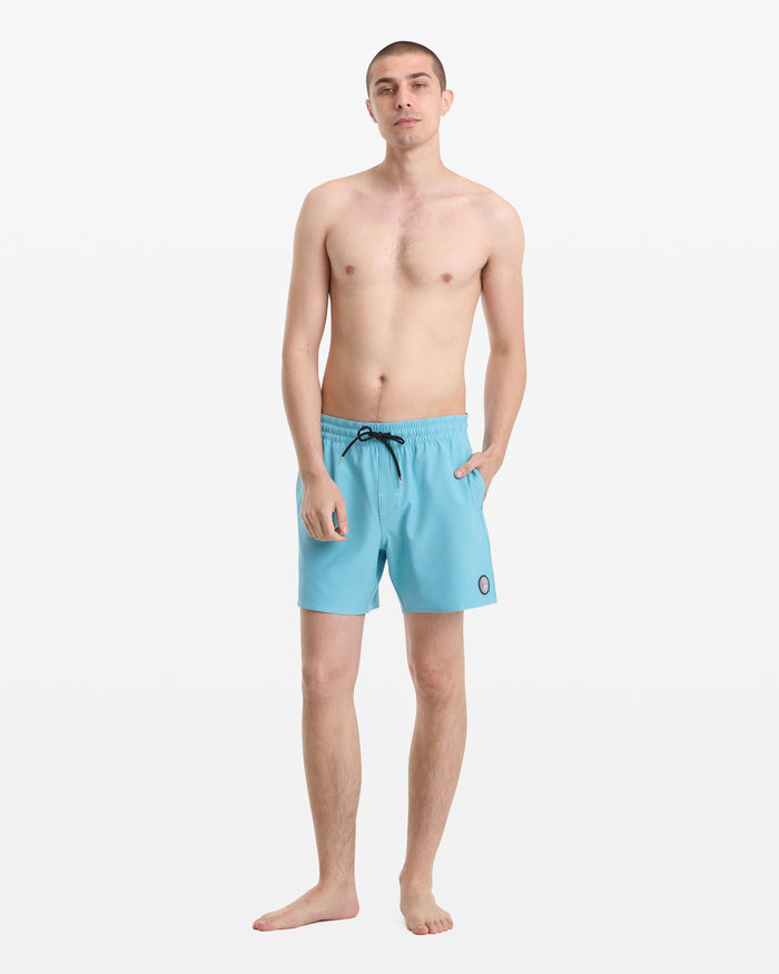 A man stands barefoot against a white background, wearing Volcom Men's Lido Solid Trunk 16 swim shorts in light blue with a black drawstring and a small round patch on the left leg. He is shirtless with short hair and his left hand in his pocket.