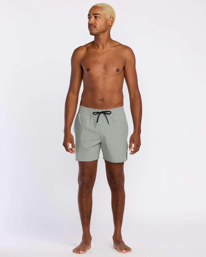 A barefoot man with short blond hair stands against a white background wearing Volcom Men's Lido Solid 16" Trunks in light gray, featuring recycled polyester, 2-way stretch, and a black drawstring. He looks slightly left in a relaxed pose.