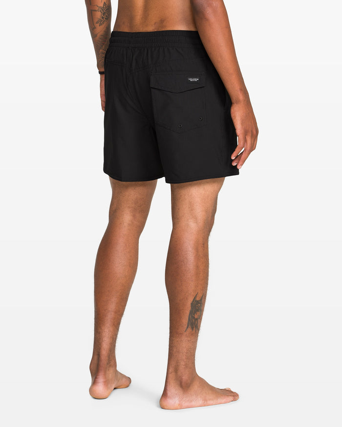 A person stands barefoot, facing away, wearing Volcom Men's Lido 2 Solid Trunks made from recycled polyester with a back pocket. Visible tattoos are on their right arm and left leg, against a plain white background.