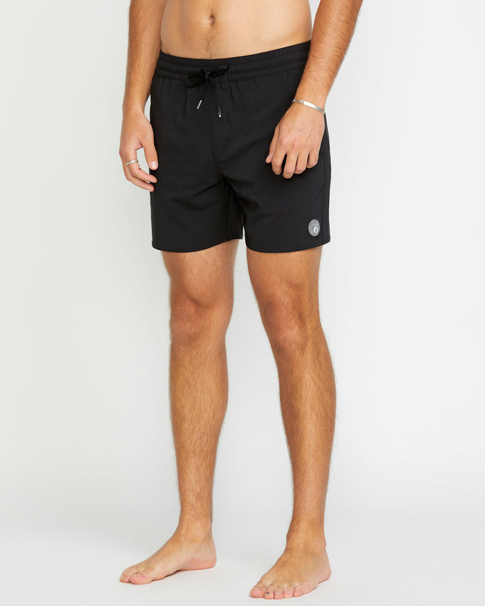 A person stands barefoot against a white background, wearing Volcom Men's Lido 2 Solid Trunks in black, featuring recycled polyester with mechanical stretch, a drawstring waist, and a small circular logo on one leg. Their upper body is not visible.