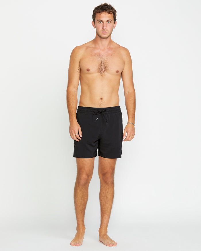A man with short brown hair stands barefoot against a white background, wearing Volcom Men's Lido 2 Solid Trunks made from recycled polyester with mechanical stretch. He faces forward with a neutral expression, arms relaxed at his sides.