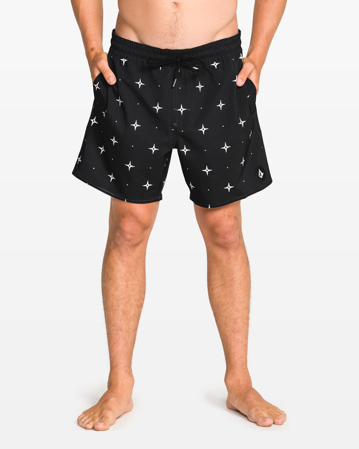 A person stands barefoot in Volcom Men's Lido Print 16" Trunk shorts with a white star print, elastic waistband with drawstring, 4-way stretch, and side pockets. The background is plain white.