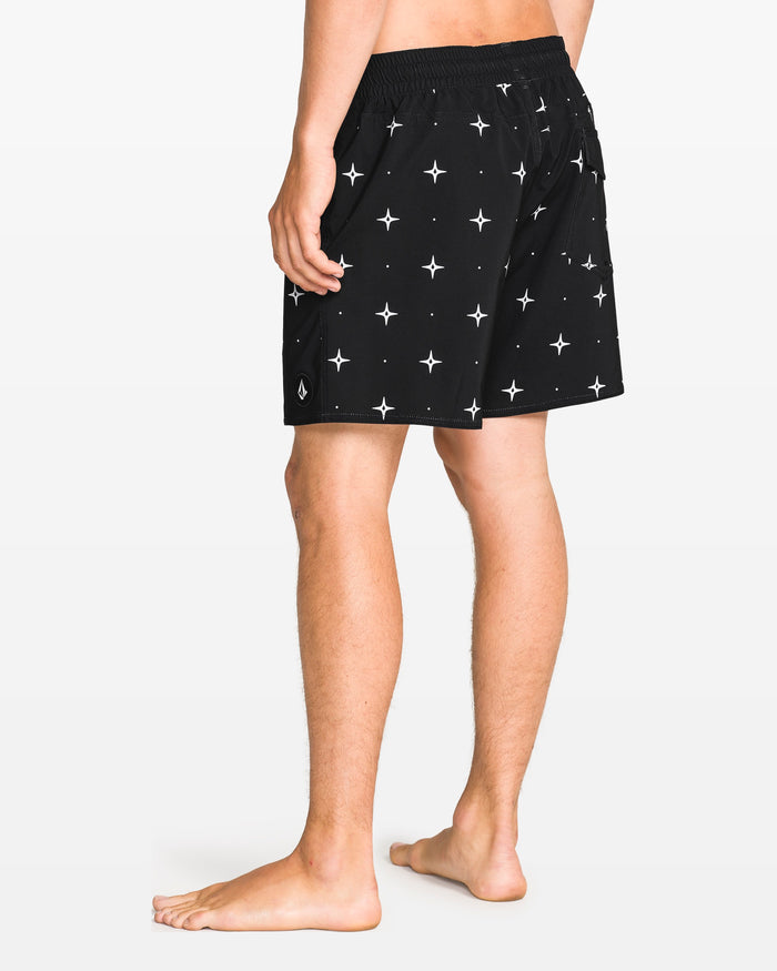 A barefoot person, shown from the waist down and facing away, wears Volcom Men's Lido Print 16" Trunk shorts in black with a white star pattern and 4-way stretch. The background is solid white.