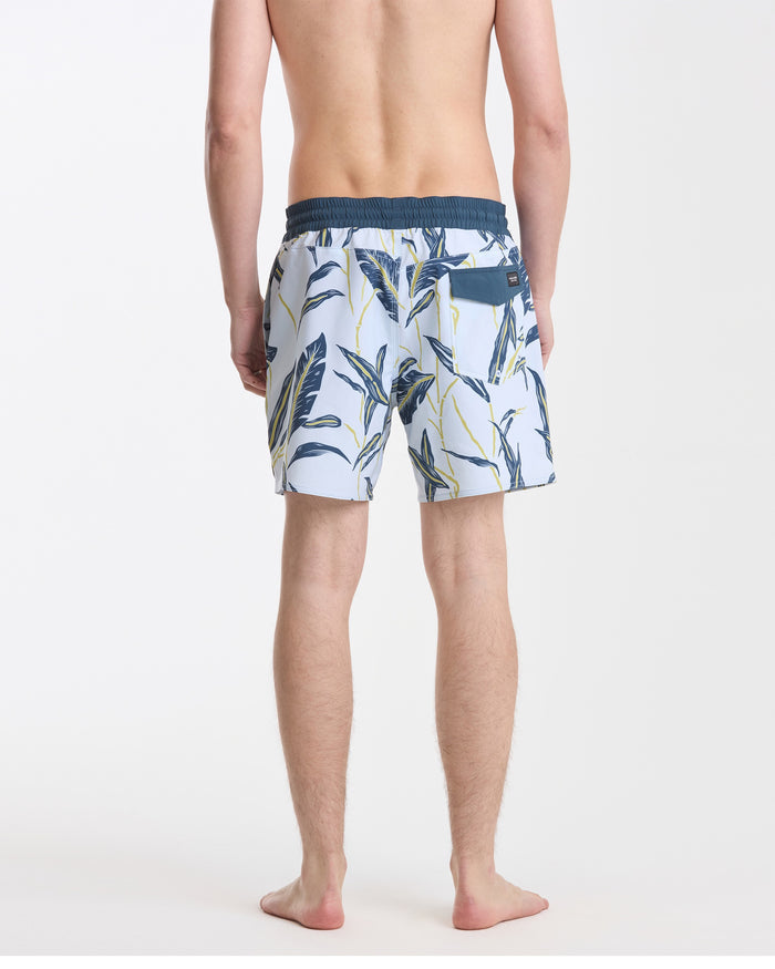A man stands barefoot facing away in Volcom Men's Lido Print 16" Trunks, white with a navy waistband and green/yellow tropical leaf pattern, made from stretchy polyester-elastane. Background is plain white.