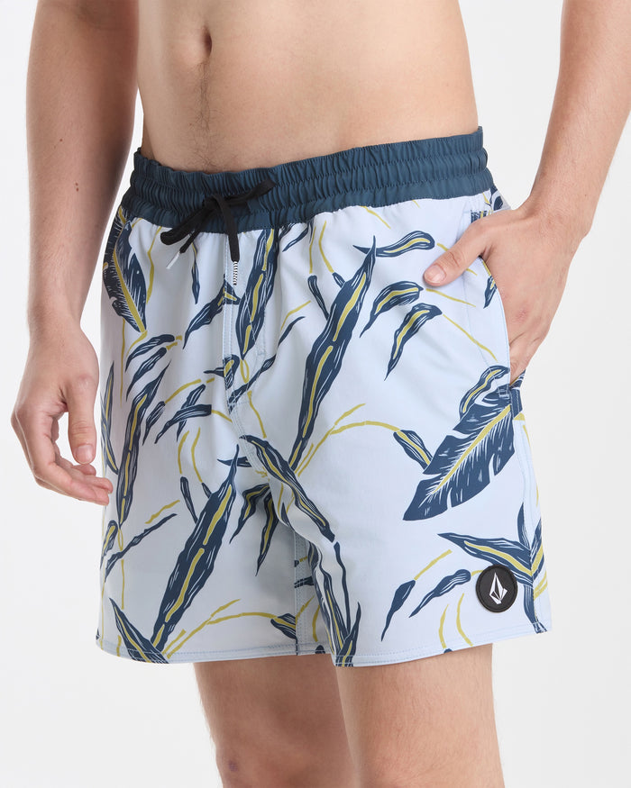 A person wears Volcom Men's Lido Print 16" Trunks—light-colored with blue and yellow palm leaf print. Made from stretchy polyester-elastane, they feature an elastic waistband, black drawstring, back pocket, logo patch, and one hand in the pocket.