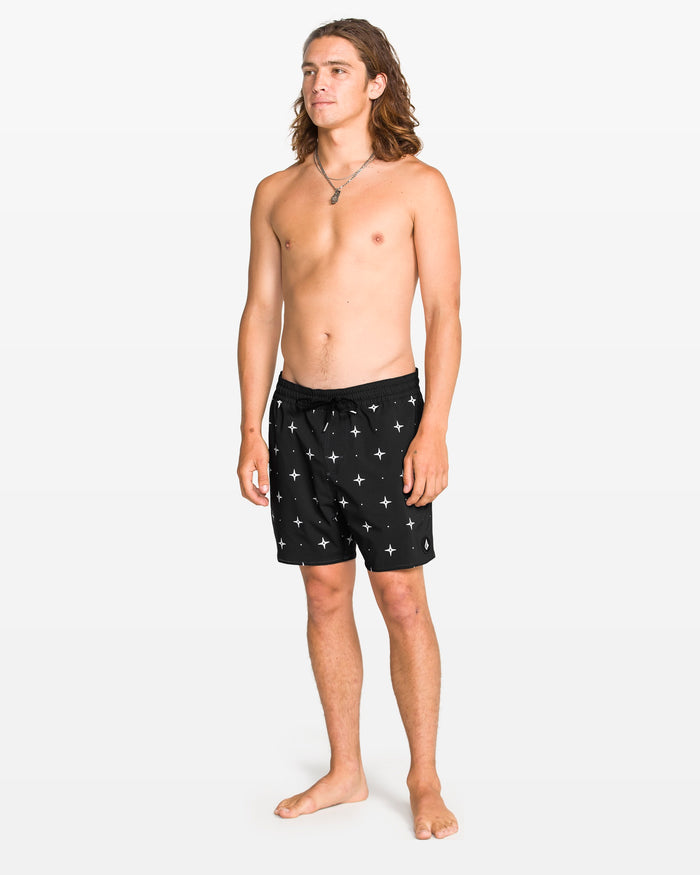 A young man with long brown hair stands barefoot on a white background, wearing Volcom Men's Lido Print 16" Trunk swim shorts with a 4-way stretch and white star pattern. He is shirtless and looks slightly to the side, appearing relaxed.