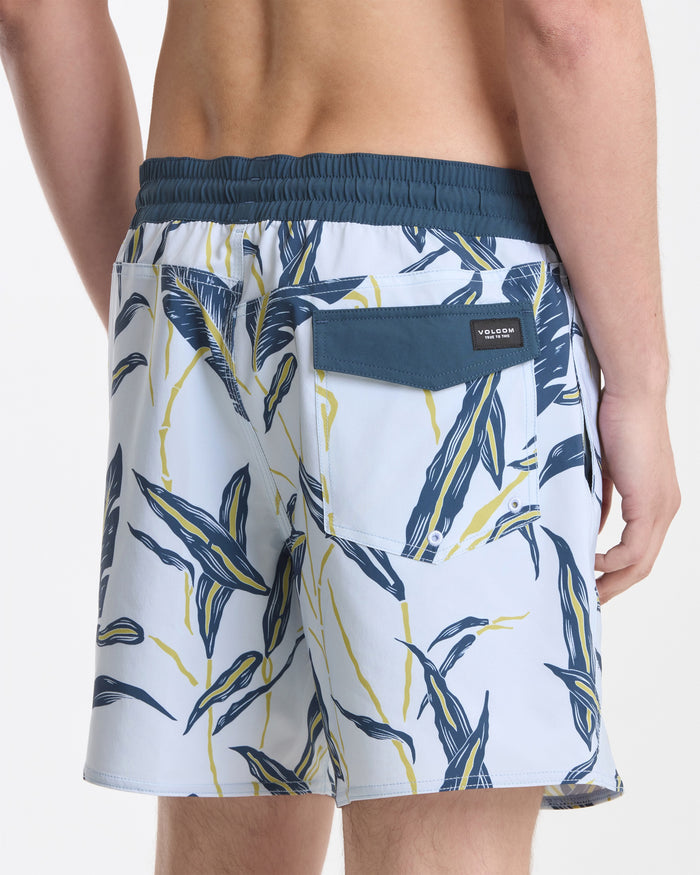 A person is shown from the waist down, viewed from the back, wearing Volcom Men's Lido Print 16" Trunks in a dark blue and yellow leaf pattern with a dark blue waistband and back flap pocket.