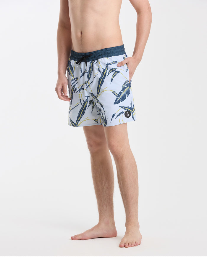 A man stands barefoot against a white background wearing Volcom’s Men's Lido Print 16" Trunks—light blue with a dark blue and yellow tropical leaf pattern, stretch polyester-elastane, dark blue waistband, back flap pocket, left hand in pocket.