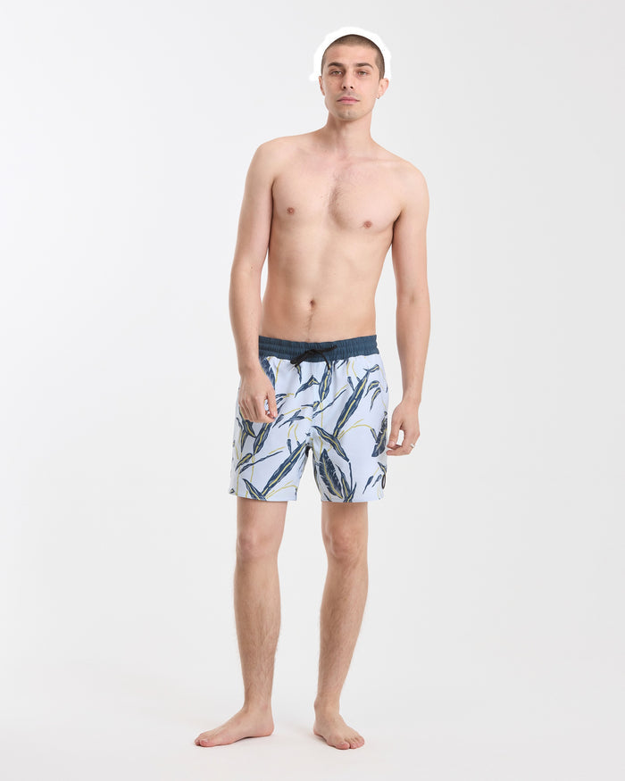 A young man stands barefoot against a plain white background, wearing Volcom Men's Lido Print 16" Trunks made from stretchy polyester-elastane with a back flap pocket, looking directly at the camera with a neutral expression.