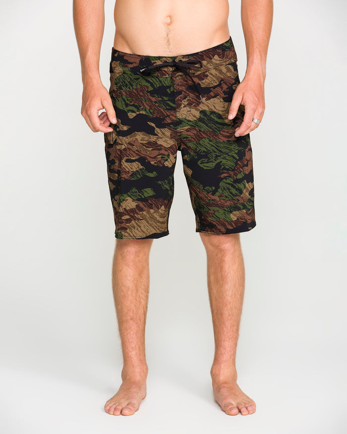A person stands barefoot wearing Volcom Men's Lido Solid Mod board shorts, crafted from recycled fibers and featuring 4-way stretch for superior comfort. Their torso is visible from the waist down against a plain, light-colored background.