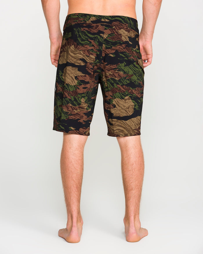 A person stands barefoot facing away, wearing green, brown, and tan knee-length camouflage shorts against a plain light background. The Volcom Men's Lido Solid Mod shorts feature a Cinch Fly and are made from 4-way stretch recycled fibers for comfort and mobility.