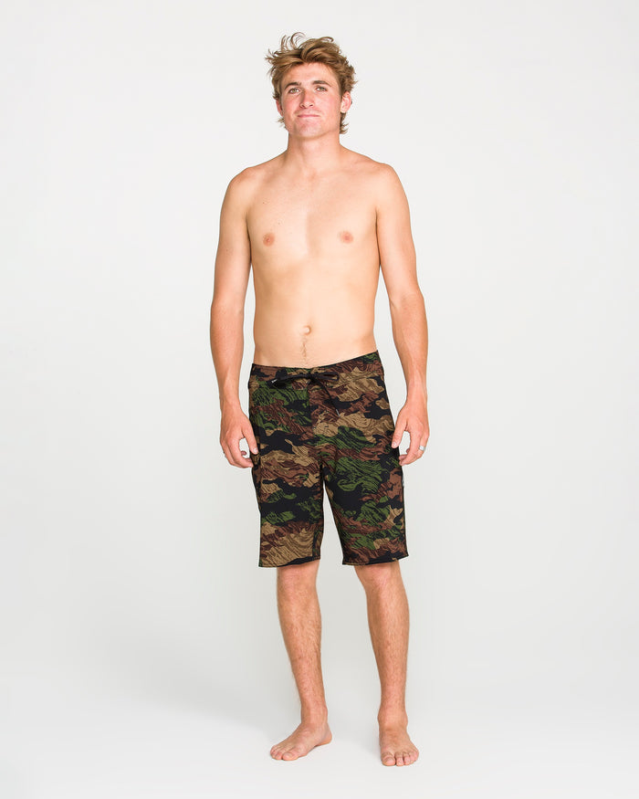 A barefoot, shirtless man with light hair stands facing forward against a plain white background, wearing Volcom Men's Lido Solid Mod shorts made from recycled fibers.