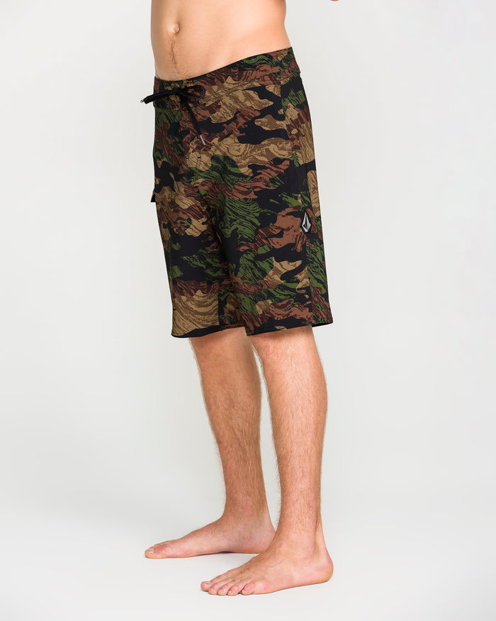 A person is standing barefoot on a plain background, wearing the Volcom Men's Lido Solid Mod shorts. Only the lower torso and legs are visible. These knee-length shorts feature a drawstring cinch fly and are crafted from 4-way stretch recycled fibers for comfort and flexibility.