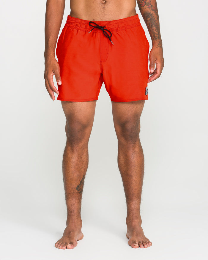 A person stands barefoot wearing bright red Volcom Men's Lido Solid Trunks, made from recycled polyester with a black drawstring and a contemporary 16-inch outseam, photographed against a plain white background.