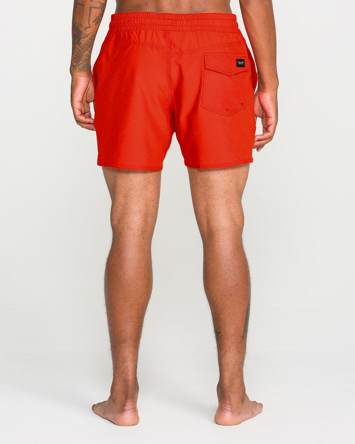 A barefoot person stands facing away, wearing Volcom Men's Lido Solid Trunks in bright red, featuring a back pocket, elastic waistband, recycled polyester construction, and a 16-inch outseam, set against a plain white background.