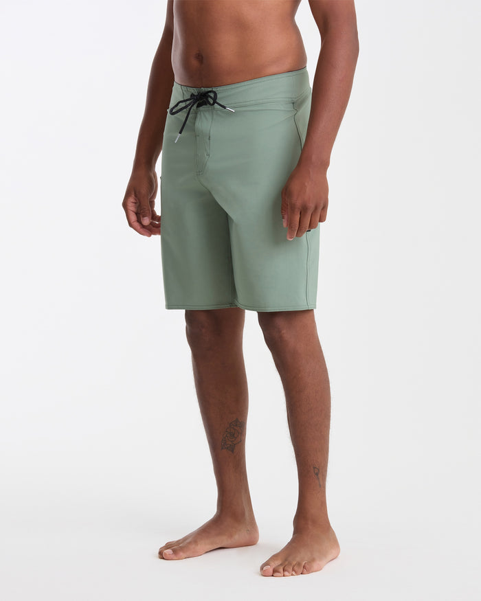 A barefoot man stands on a white background, wearing Volcom Men's Lido Solid 20" Trunks in light green with a black drawstring. His torso is bare and he has a small tattoo on his left leg.