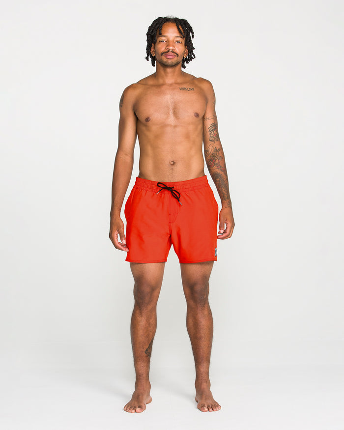 A man with short dreadlocks stands facing forward, shirtless and displaying tattoos on his chest and left arm, against a plain white background. He is wearing Volcom Men's Lido Solid Trunks in a vibrant red color with a 16-inch outseam. These trunks are crafted from recycled polyester, combining sustainability with style.