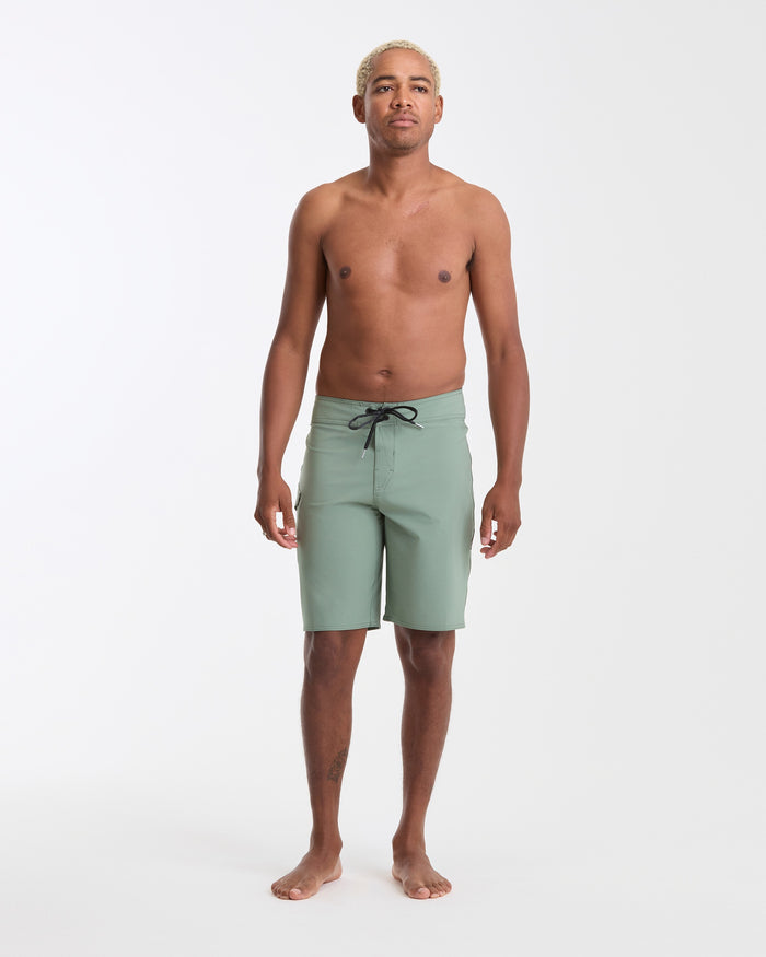 A man with short, bleached hair stands barefoot against a plain white background, wearing Volcom Men's Lido Solid 20" Trunks in light green made from recycled polyester. His arms are relaxed at his sides.