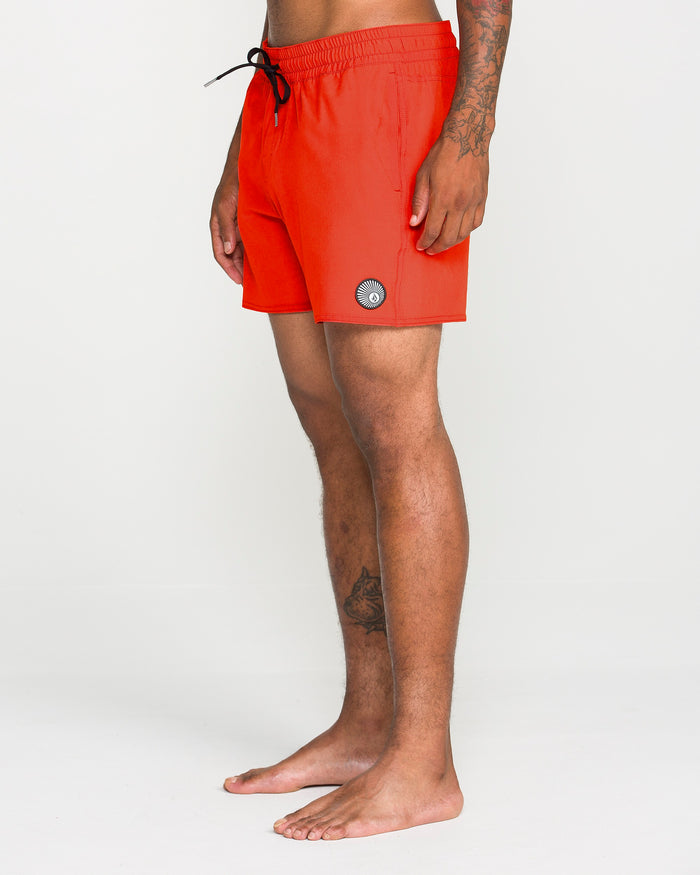 A person stands barefoot against a plain light background, wearing Volcom Men's Lido Solid Trunks in bright orange with a 16-inch outseam, featuring a black drawstring and a small circular patch on the left leg, crafted from recycled polyester.